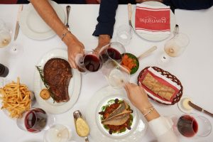 An elegantly set table in a French brasserie, with several people, friends or family, sharing a convivial meal. The main dish is a côte de bœuf, accompanied by golden fries and béarnaise sauce. There is a basket of artisanal bread, vegetables (carrots and peas), and several glasses of red wine to complement the côte de bœuf. Three people are toasting with wine, illustrating a warm, convivial, and festive atmosphere. The Brasserie Gavroche logo is visible on a red napkin, reinforcing the restaurant's identity. The entire scene evokes a refined dinner, authentic French cuisine, and a warm, welcoming ambiance, perfect for meals with friends, business lunches, or group dinners.